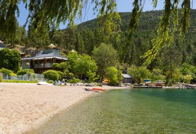 A serene lakeshore with a building, trees, and boats on a sunny day.