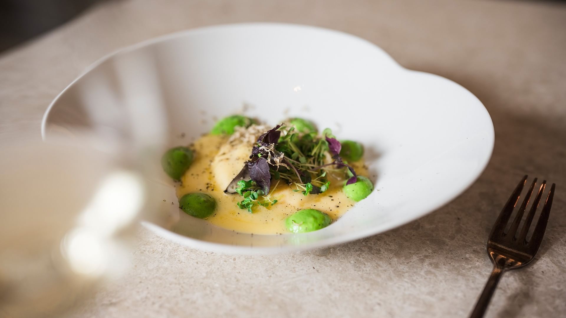 Weiße Schüssel mit grünen Erbsen, gelber Sauce und Kräutern, daneben eine Gabel auf einer Tischoberfläche.