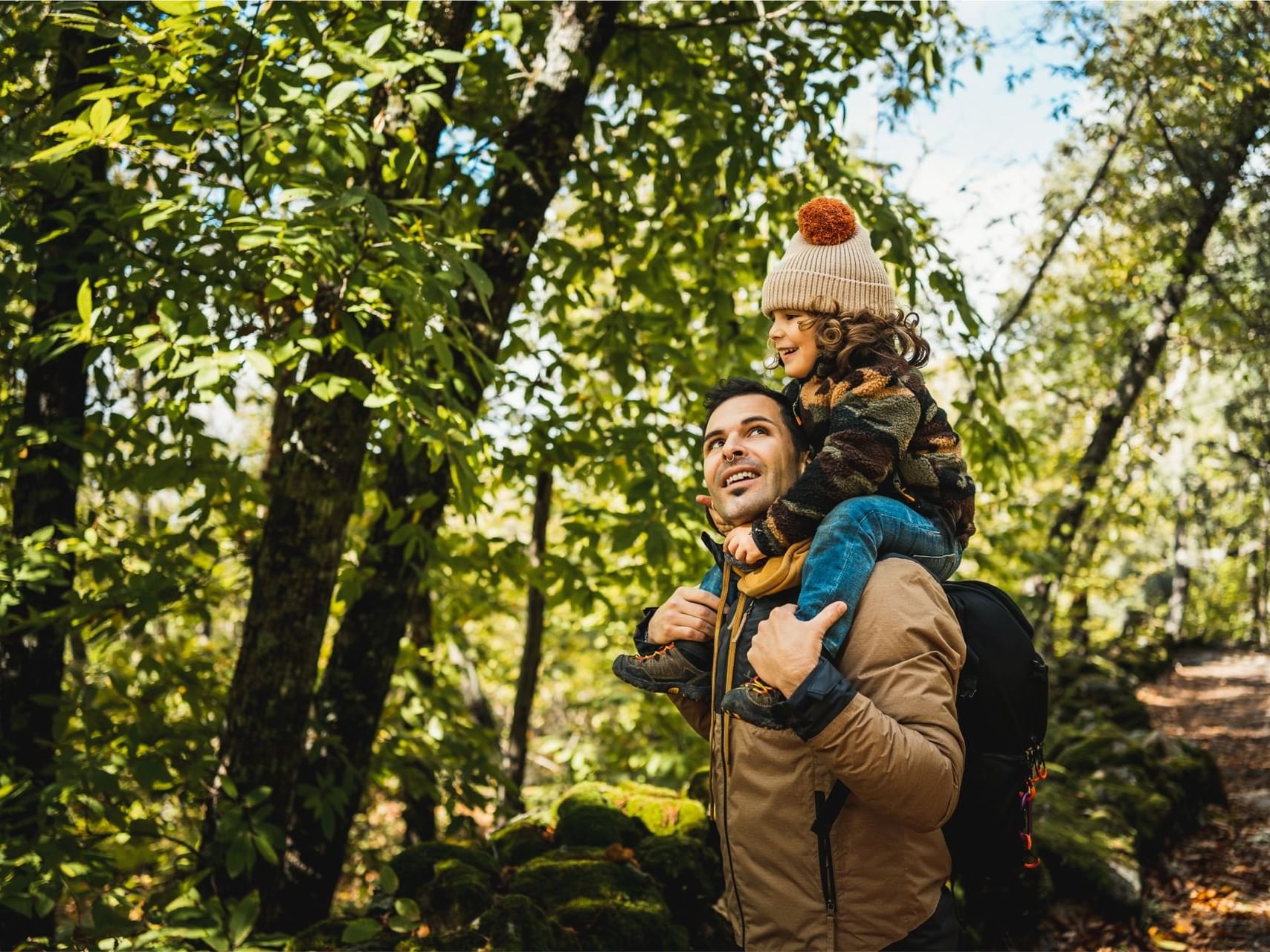 Father carrying a young child on his shoulders while hiking in a green, lush forest near Fall Creek Marina & Campground