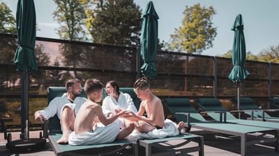 Family of four in robes relaxing on lounge chairs by a pool with closed umbrellas.
