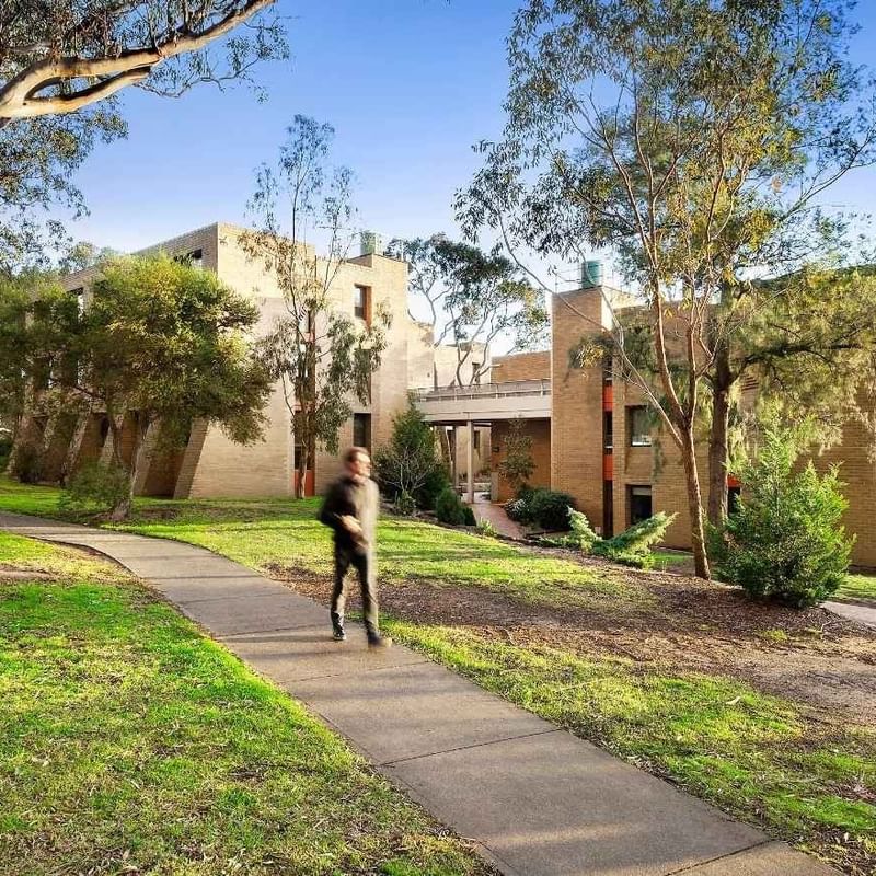 Person walking on sidewalk towards modern buildings and lush greenery at La Trobe University - Chisholm College.