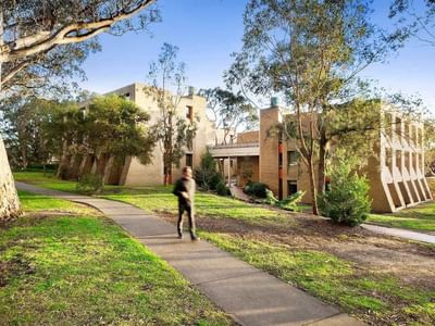 Person walking on sidewalk towards modern buildings and lush greenery at La Trobe University - Chisholm College.
