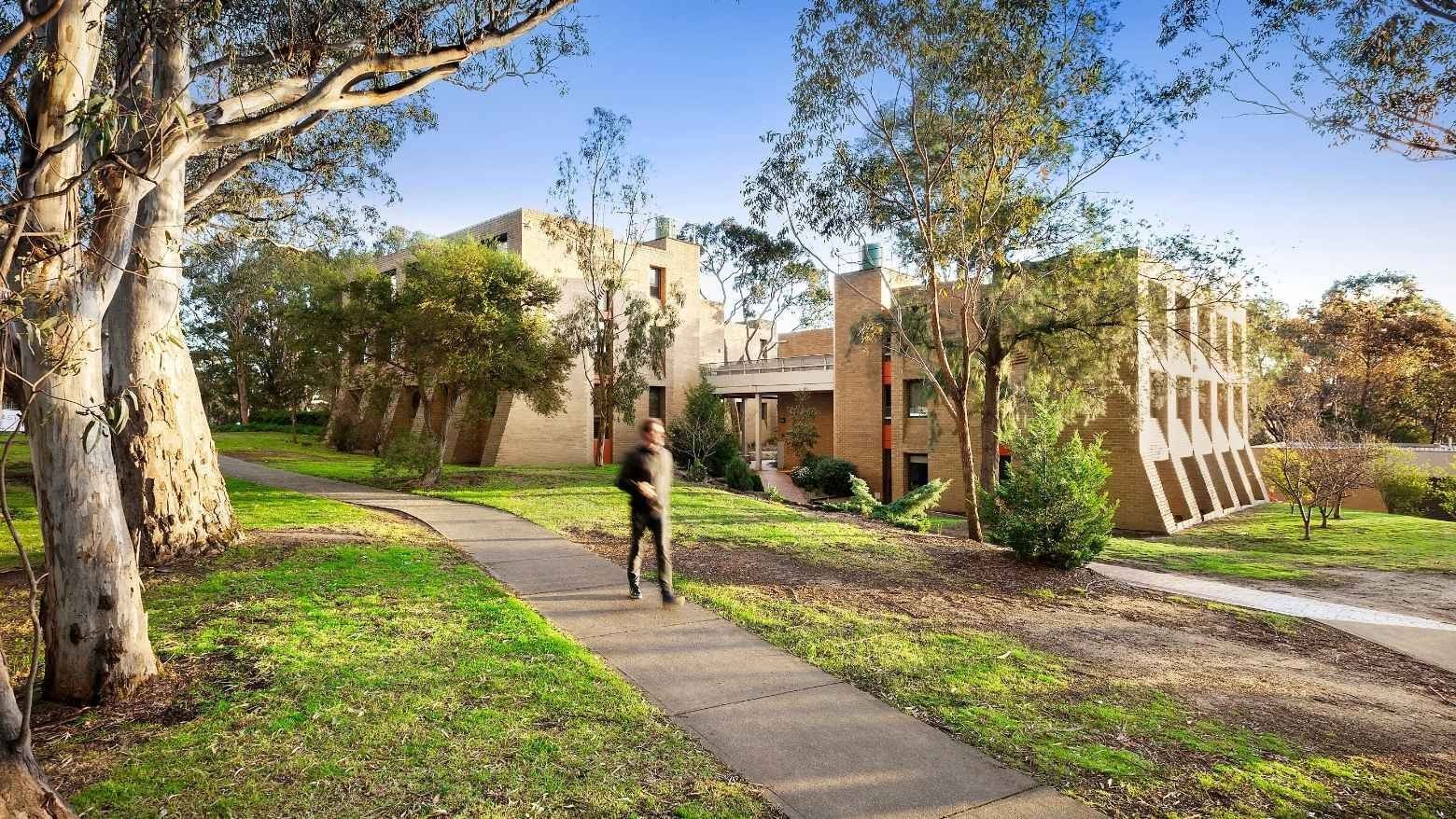 Person walking on sidewalk towards modern buildings and lush greenery at La Trobe University - Chisholm College.