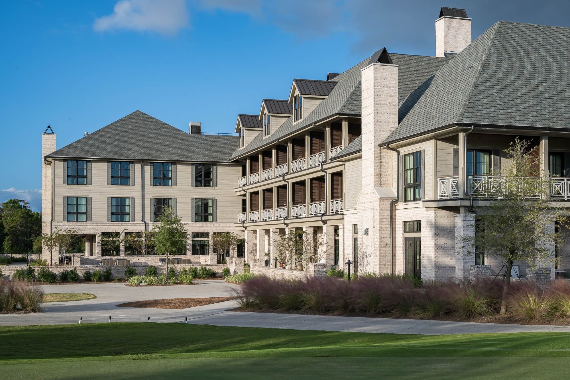 Modern building with balconies and gray roof, surrounded by greenery and grass under a blue sky.
