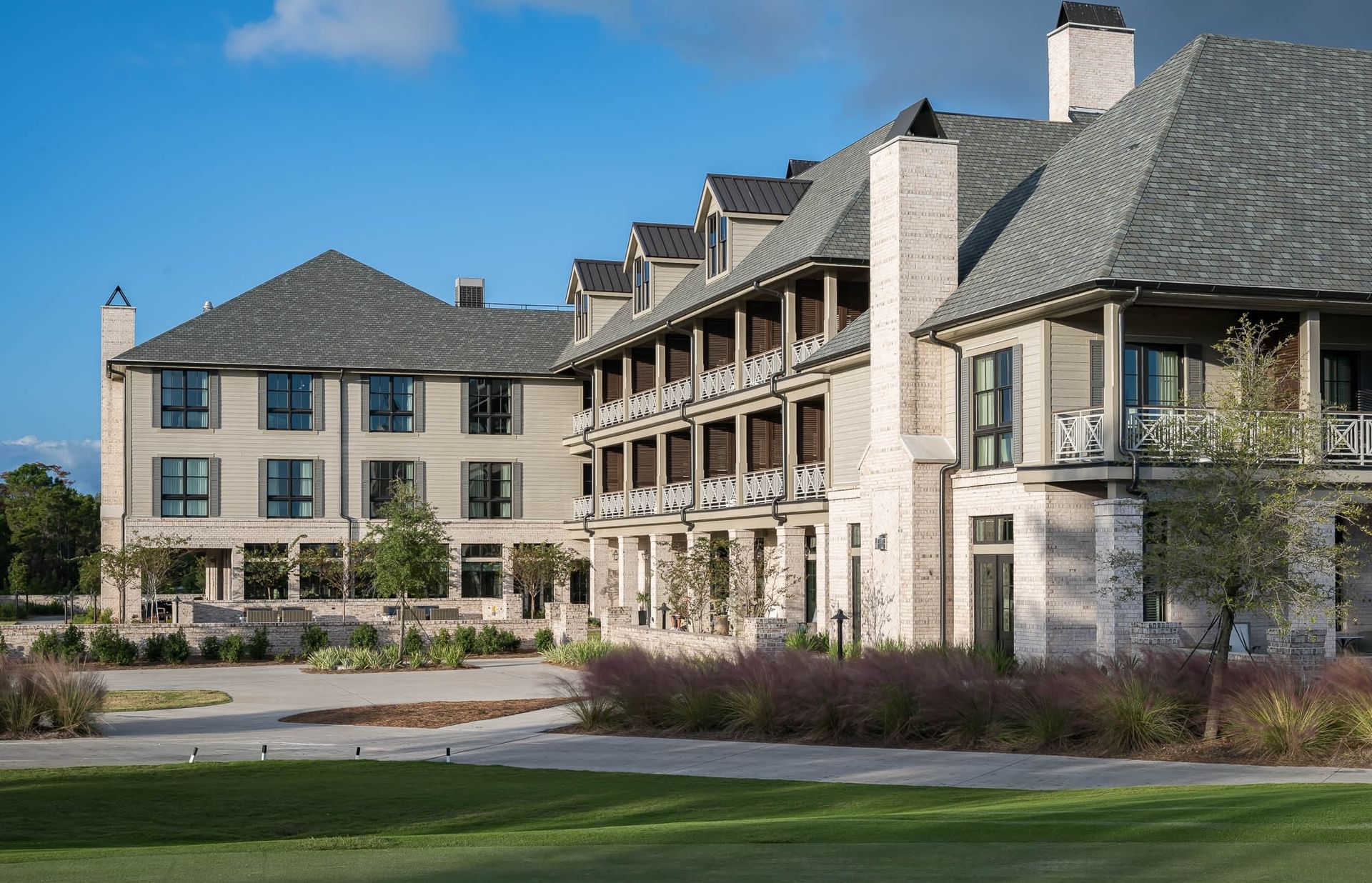 Modern building with balconies and gray roof, surrounded by greenery and grass under a blue sky.