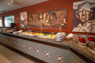 Long breakfast buffet counter under a protective sneeze guard by a painted mural wall in Fonda de San Luis at Hotel Mirra