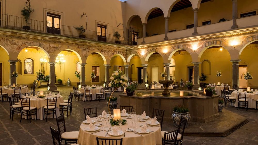 Evening outdoor banquet in a stone courtyard with white clothed tables and a fountain in Patio Novicias at Quinta Real Puebla