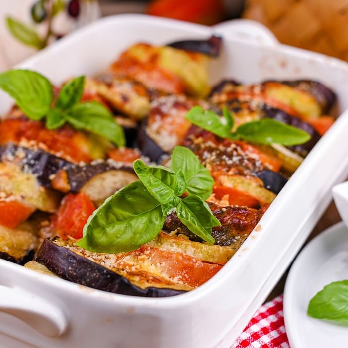 Gebackene Auberginen mit Tomaten und Basilikum in einer weißen Auflaufform, begleitet von einem Teller mit Basilikum.