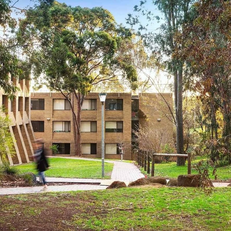 Student walking toward La Trobe University - Chisholm College building in a lush green park.
