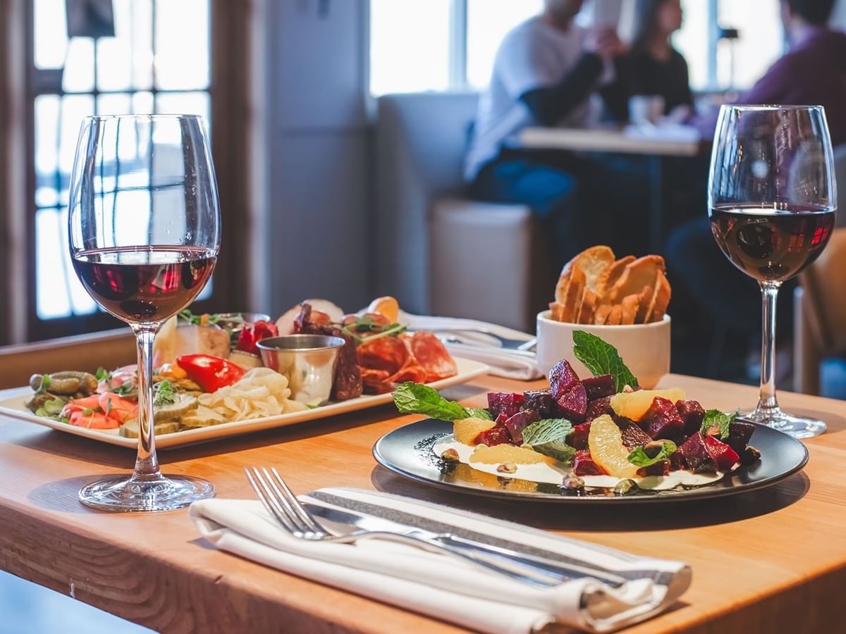 A table with two plates of food, two wine glasses, a bowl of bread, and cutlery.