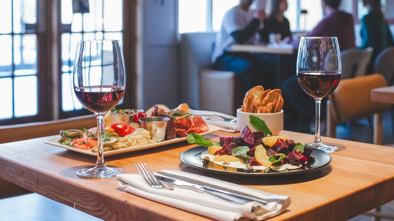 A table with two plates of food, two wine glasses, a bowl of bread, and cutlery.