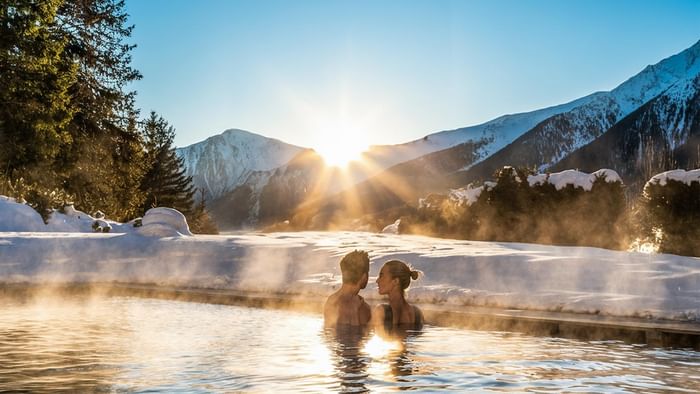 Paar entspannt im Pool mit schneebedeckten Bergen bei Sonnenuntergang am Falkensteiner Group