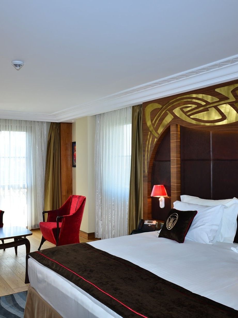 Red chairs by a coffee table surrounding a plush bed under a gold headboard in Junior Suite at Warwick Ankara
