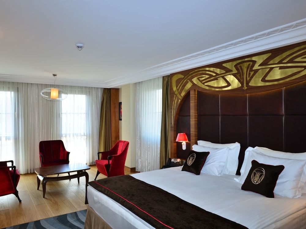 Red chairs by a coffee table surrounding a plush bed under a gold headboard in Junior Suite at Warwick Ankara