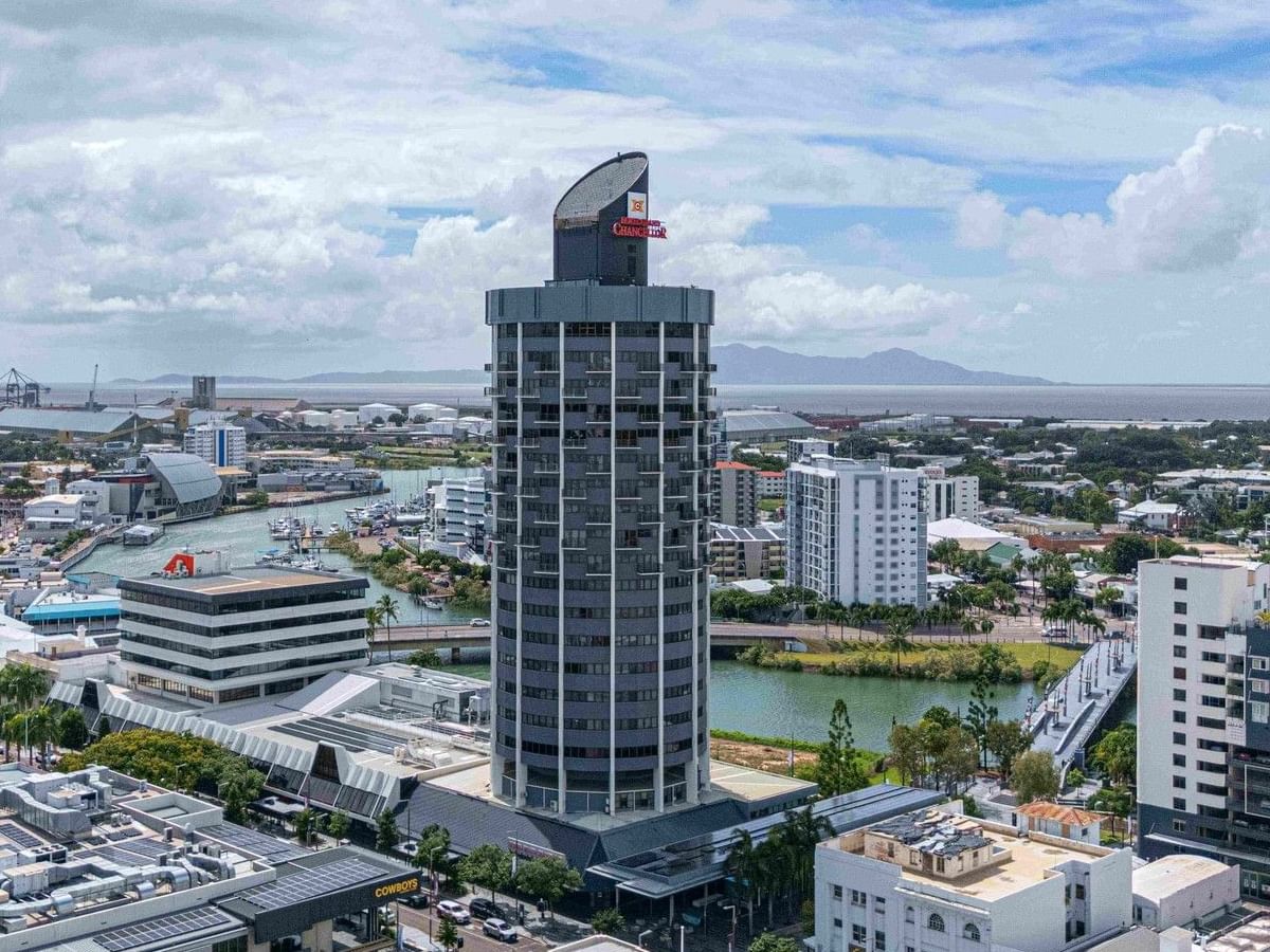 Aerial view of Hotel Grand Chancellor Townsville, a distinctive high-rise tower overlooking the Ross Creek waterfront, with the city centre, marina, port facilities, and distant mountains under a partly cloudy sky.