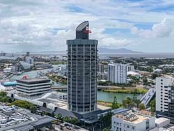 Aerial view of Hotel Grand Chancellor Townsville, a distinctive high-rise tower overlooking the Ross Creek waterfront, with the city centre, marina, port facilities, and distant mountains under a partly cloudy sky.