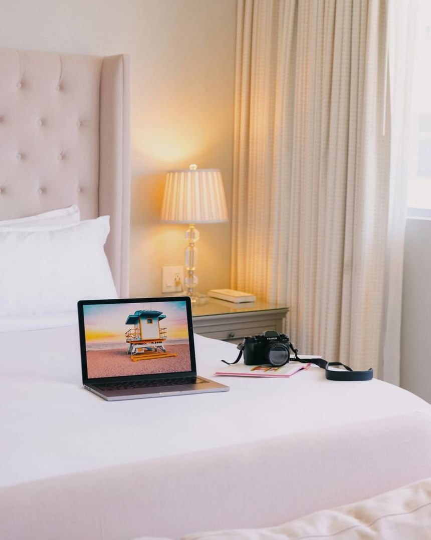 Laptop displaying a beach photo and a camera resting on a plush white bed in a cozy room at The Plymouth Hotel South Beach