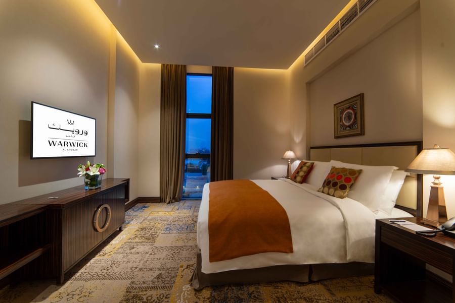 King-sized bed by a dark wood dresser under a glowing ceiling cove in Business Suite at Warwick Al Khobar