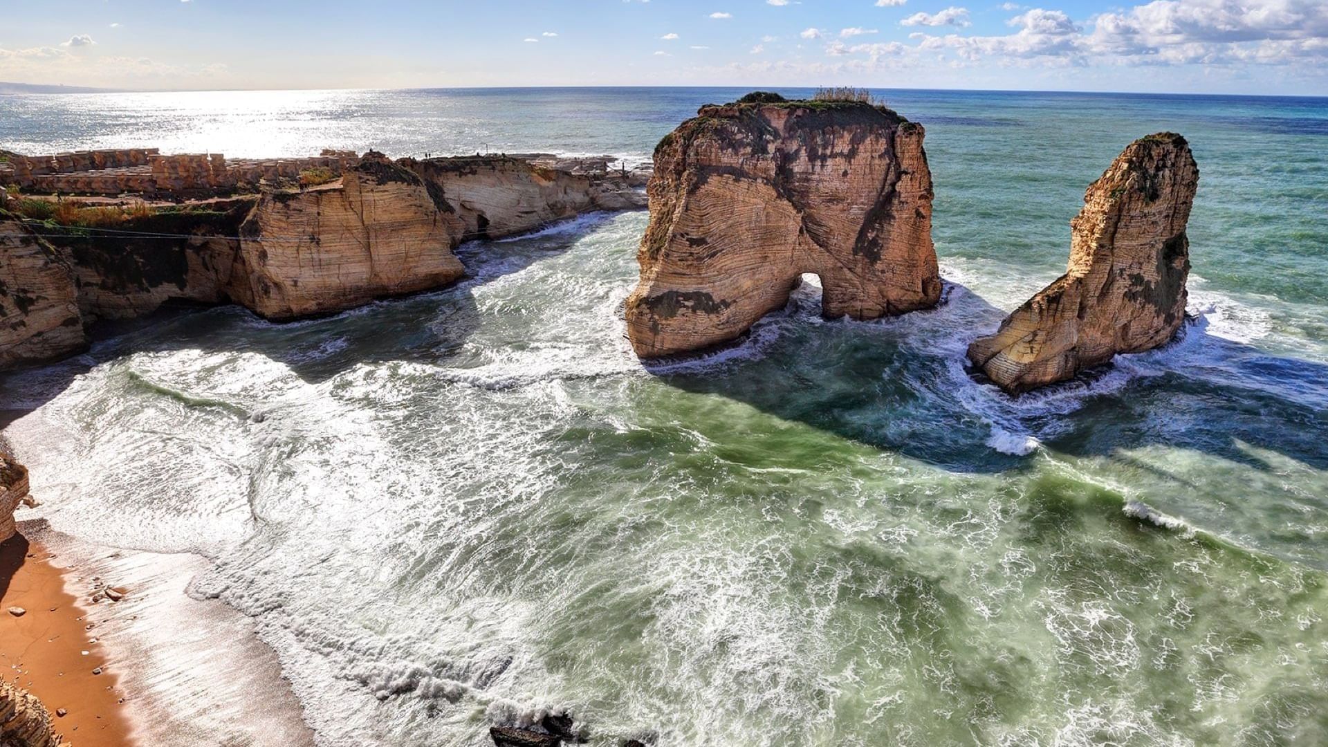Large rock arches in the sea by a sandy beach under a cloudy blue sky near the Warwick Palm Beach Hotel