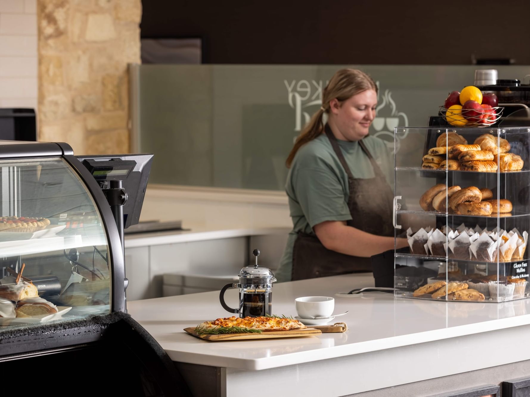 Server preparing coffee at station, with flatbread and coffee on the counter by the coffee shop display case at Monkey Grind Coffee Shop at Shangri-La Resort and Golf Club