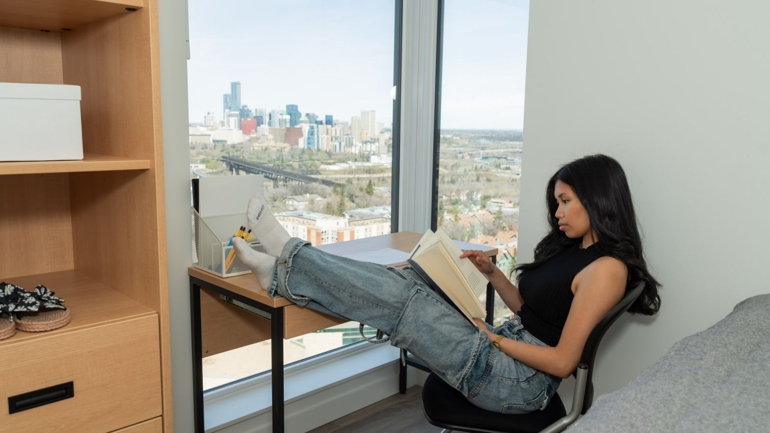 A student comfortably reads at a desk in a bedroom with views of a sunny downtown skyline in the distance.