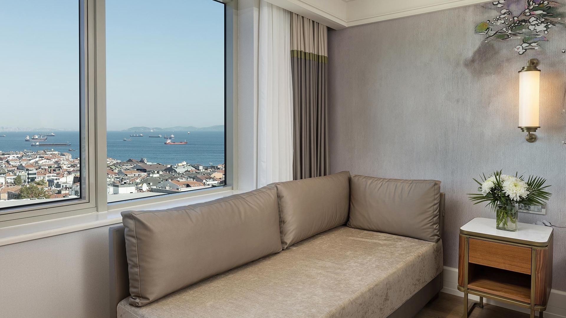 Deluxe Sea View Room with a couch, a side table, a flower vase, and a view of Marmara sea through a large window in Titanic Port Bakırköy.