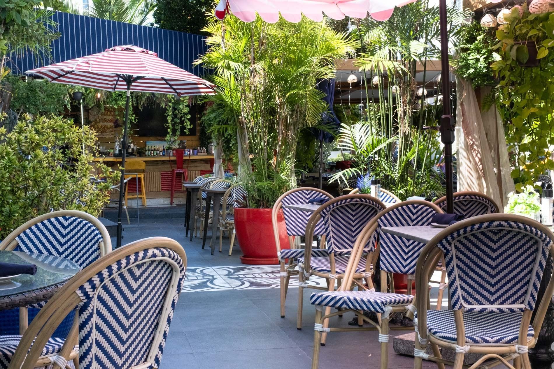 An outdoor dining area with blue and white chairs, red and white umbrellas, and potted plants.