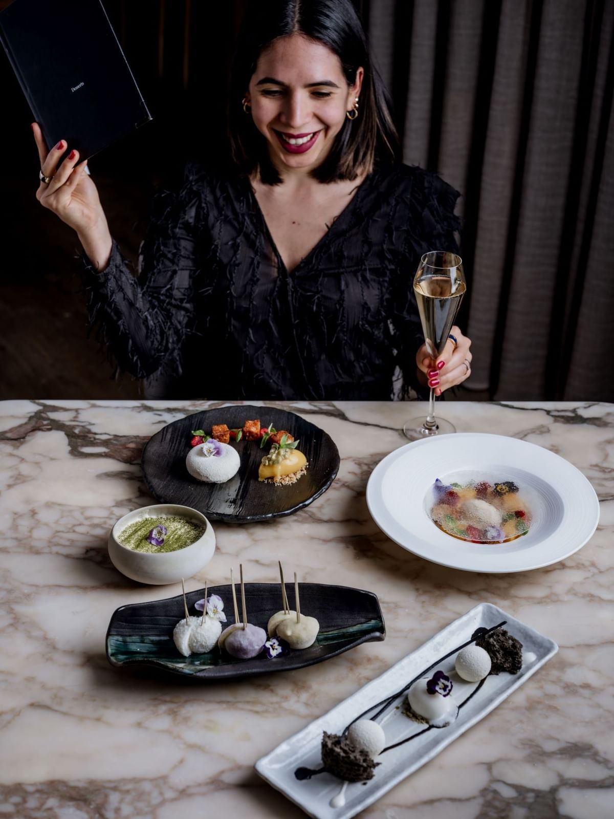 Woman in black dress smiles holding a champagne flute with elegant desserts on a marble table at The May Fair Hotel