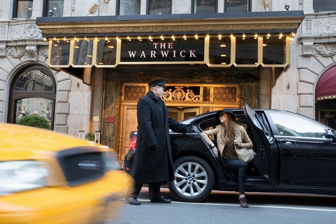 Hotel doorman holds a black sedan door open for a female guest under a lit canopy at Warwick New York