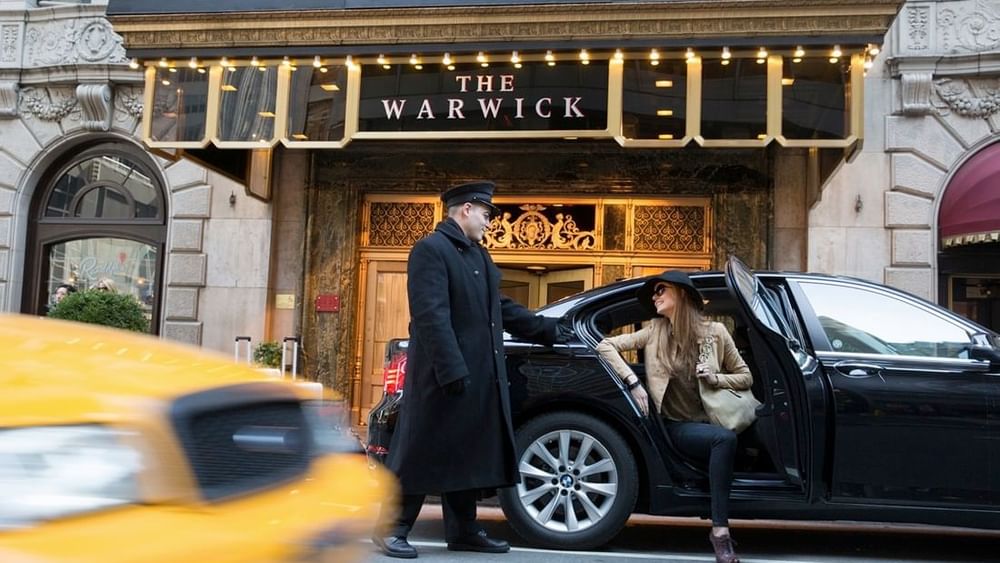 Woman exiting a car by a doorman, with a yellow taxi driving by the entrance at Warwick Hotels & Resorts