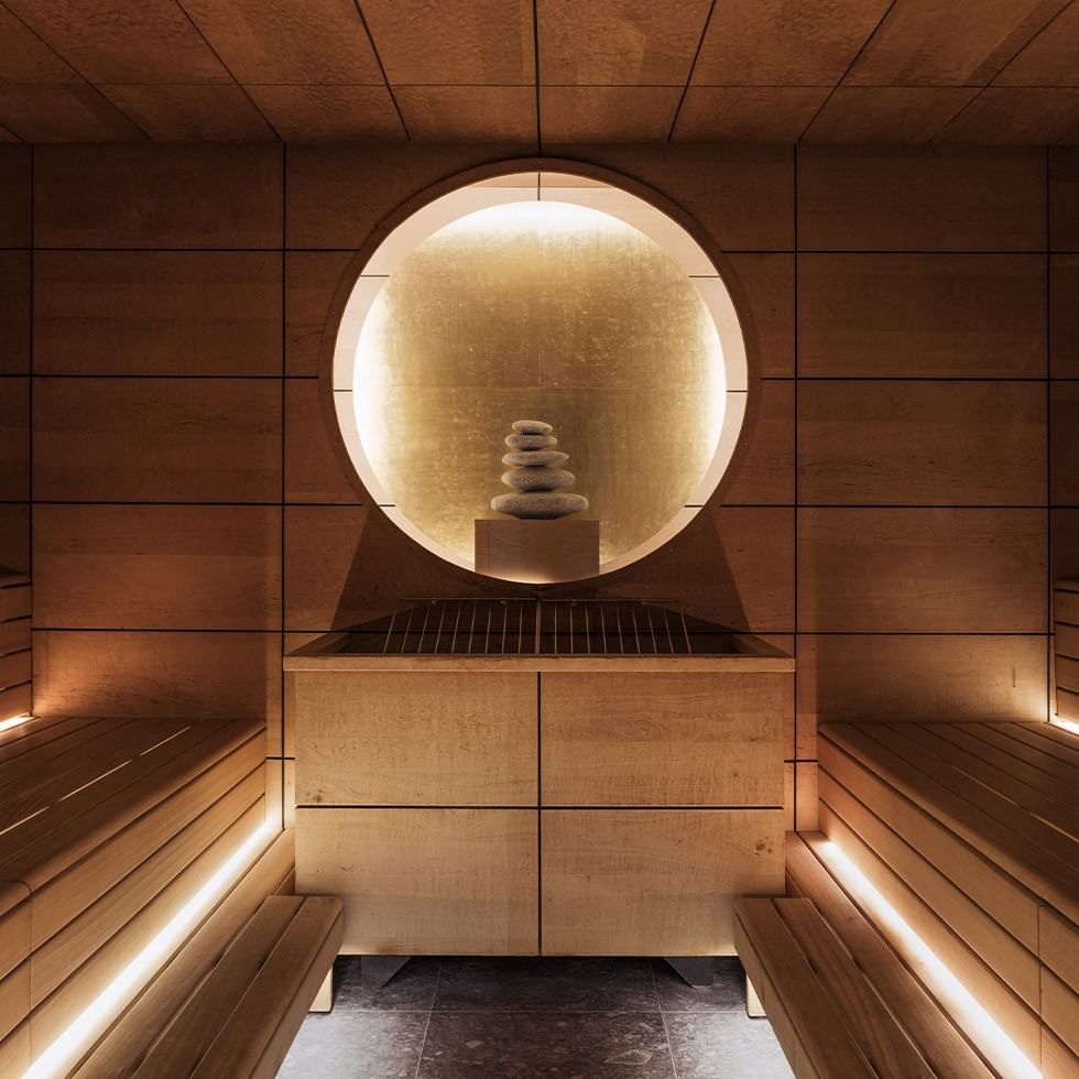 Sauna room with wooden benches and a glowing circular window above a stone structure.