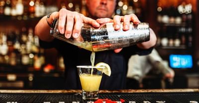 Bartender pouring a cocktail from a shaker into a shot glass with a lemon wedge.