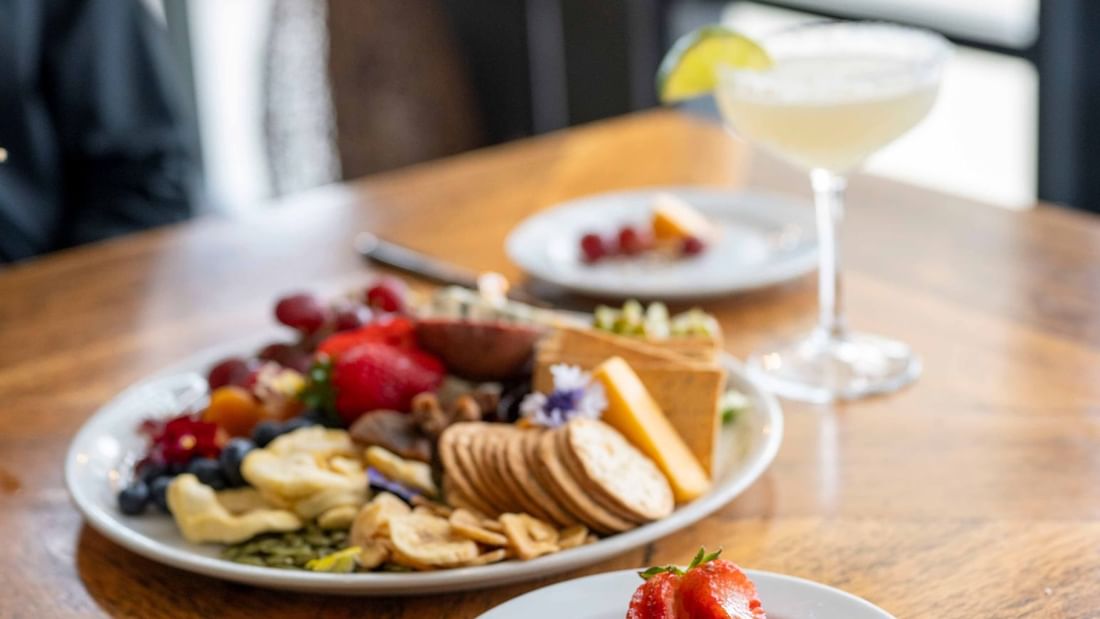 Two plates of assorted food, a cocktail, and a glass on a wooden table.