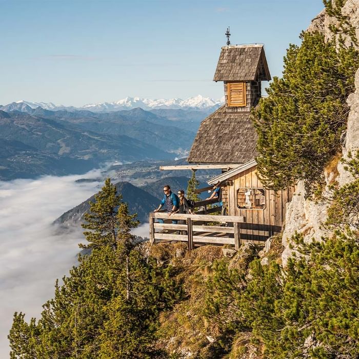 Three people on a balcony by a mountain cabin with a view of distant peaks and valleys.