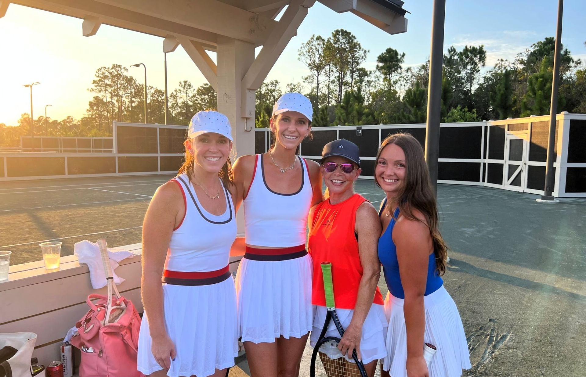 Four women in tennis outfits pose together on a tennis court with a sunset in the background.