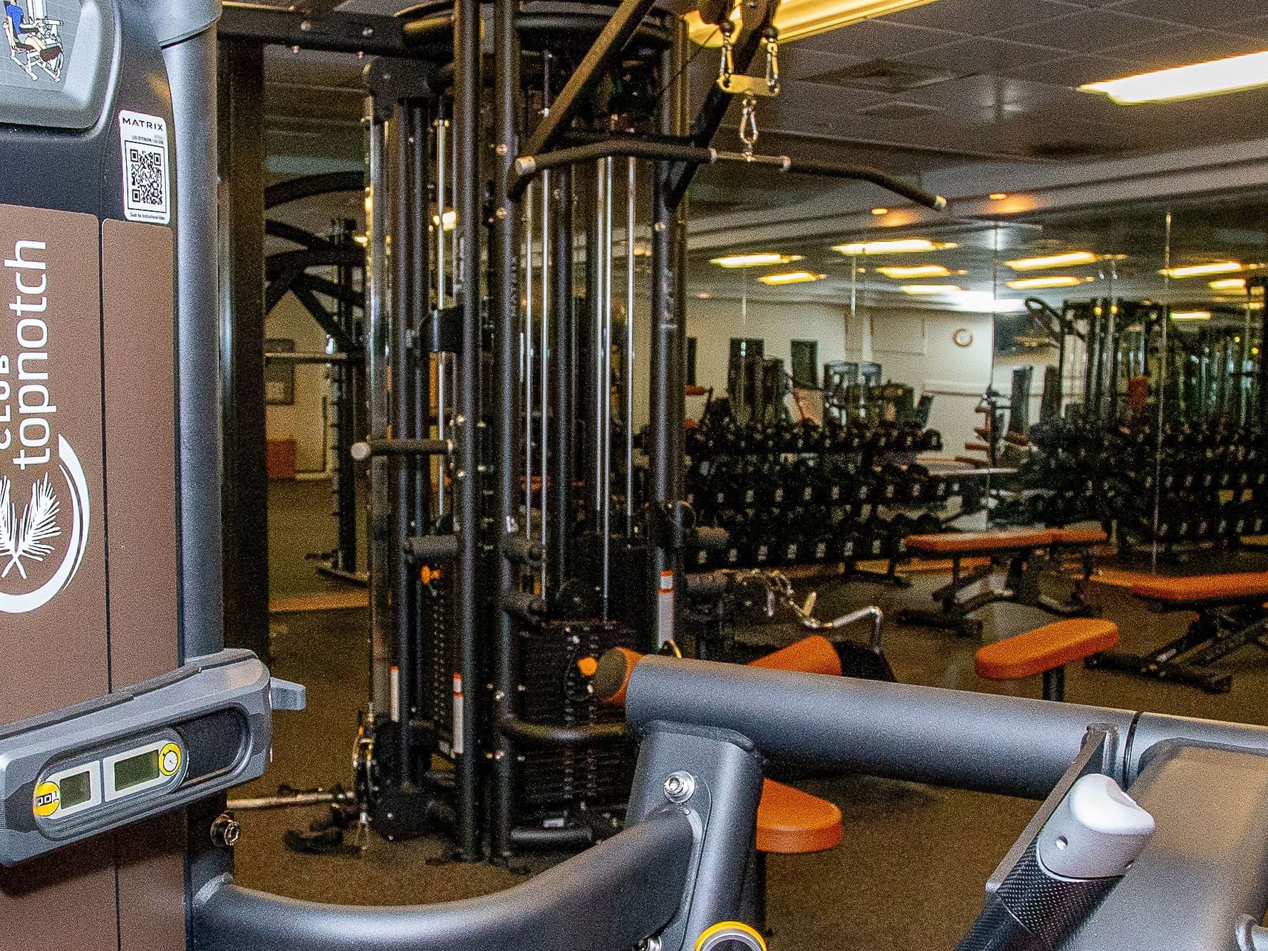 Interior of a gym with exercise machines and free weights, under bright ceiling lights and orange benches.