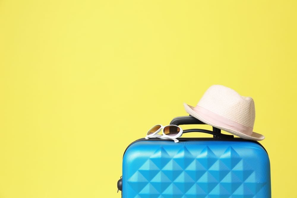 The top of a bright blue suitcase with a straw hat and pair of white sunglasses against a bright yellow background.