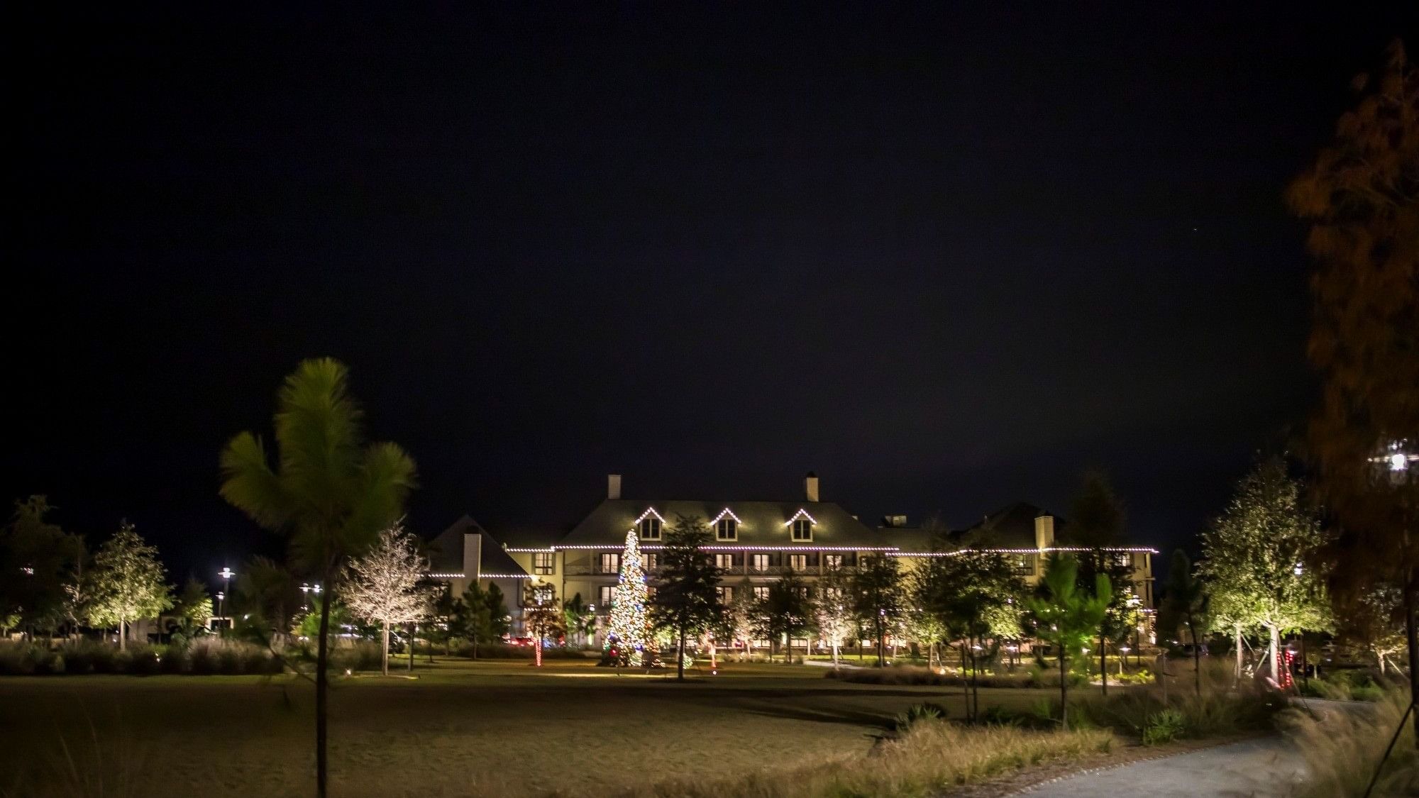 Nighttime view of a large, illuminated hotel with festive lights and trees, surrounded by a grassy area and pathway.
