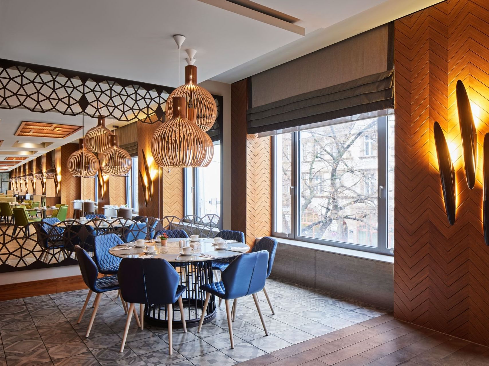 Elegantes Restaurant mit runden Tischen, blauen Stühlen, hölzernen Pendelleuchten und großen Fenstern – Titanic Chaussee Berlin.