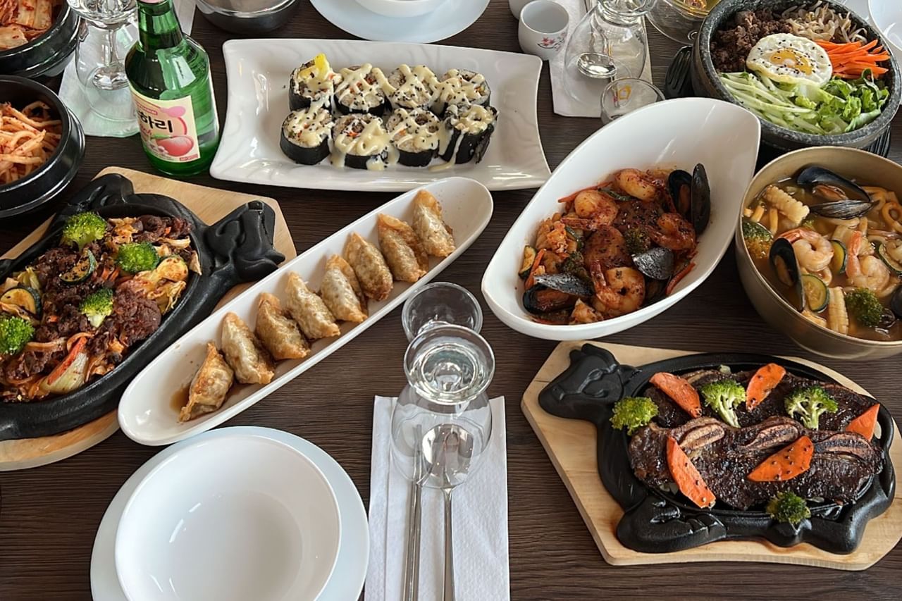 An array of Asian dishes on a wooden table with plates, bowls, and a bottle of drink.