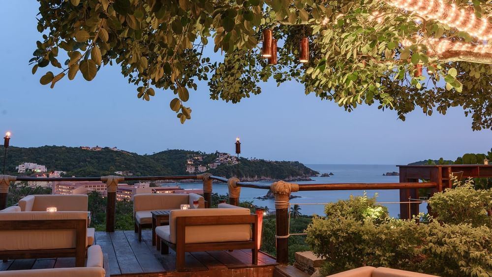 Sky Lounge area featuring cream sofas by a wood railing under a leafy canopy at night at Quinta Real Huatulco