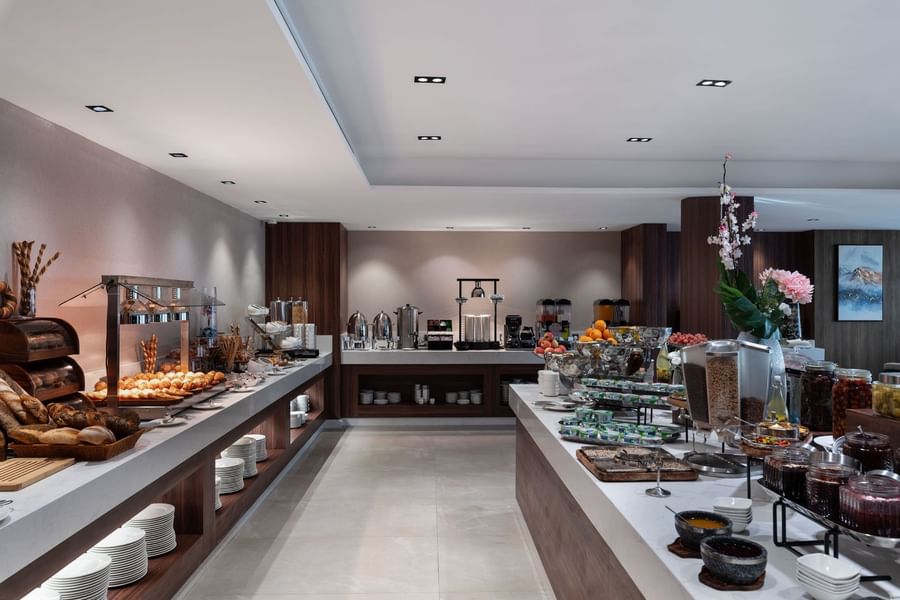 Wide buffet with pastries & cereals by wooden panels under bright spotlights in Rosa Restaurant at Warwick Al Taif Hotel