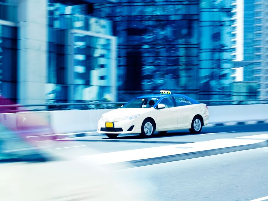 A white Dubai Taxi in motion blur speeding along a street near DAMAC Hotels & Resorts