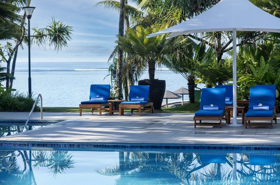Poolside deck with blue chairs by a white umbrella under palm trees at Warwick Fiji Resort and Spa