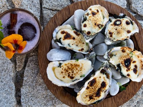Plate of baked oysters on stones, next to a festive cocktail in De Cortez Grill House at Hacienda del Mar los Cabos