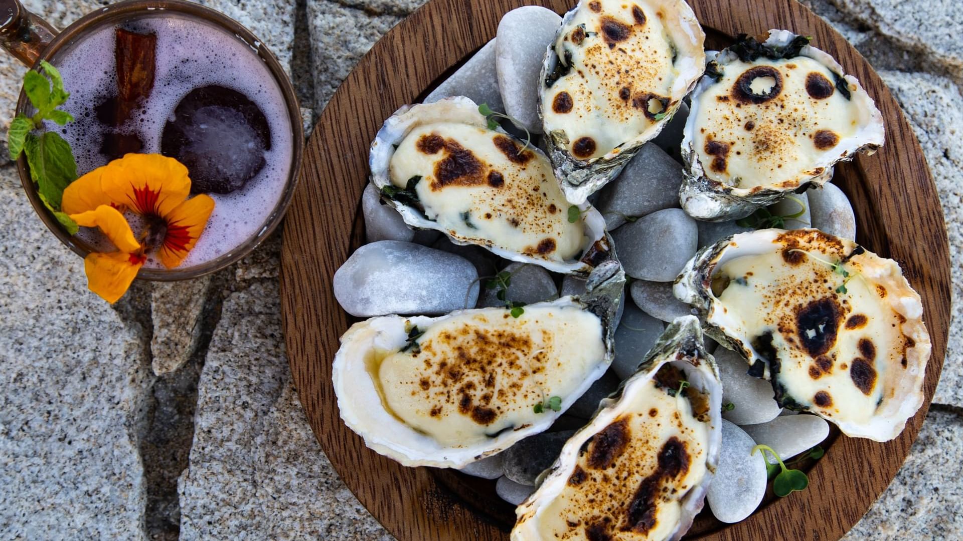 Plato de ostiones horneados sobre piedras, junto a un cóctel festivo en De Cortez Grill House de Hacienda del Mar Los Cabos