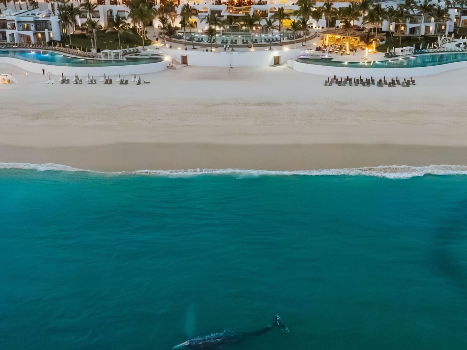 Vista aérea de resort frente al mar con playa de arena blanca, piscinas y montañas al fondo al atardecer