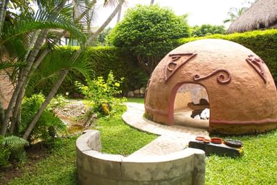Clay dome structure with carved designs in a garden with greenery, trees, and a stone path.