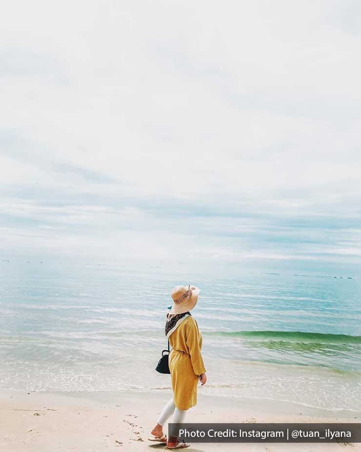 A person in a yellow coat standing by a calm beach, gazing at the sea - Lexis Port Dickson