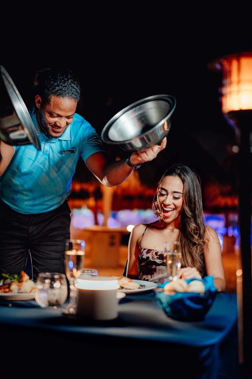 Waiter showing food to a smiling woman at a table set with wine and bread.
